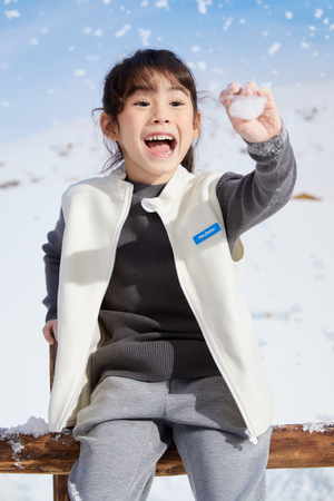 A girl in a gray sweater and white soft lightweight vest holding a snowball in a snowy landscape
