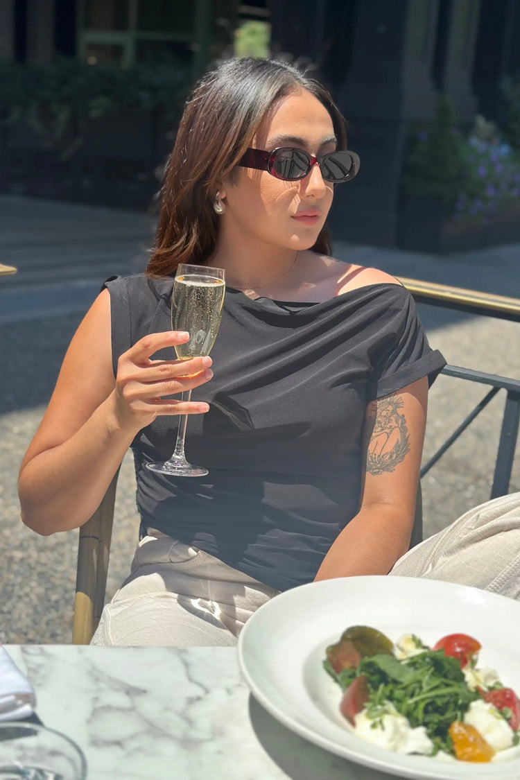 Women’s asymmetric draped top worn with sunglasses and linen pants, holding a champagne glass