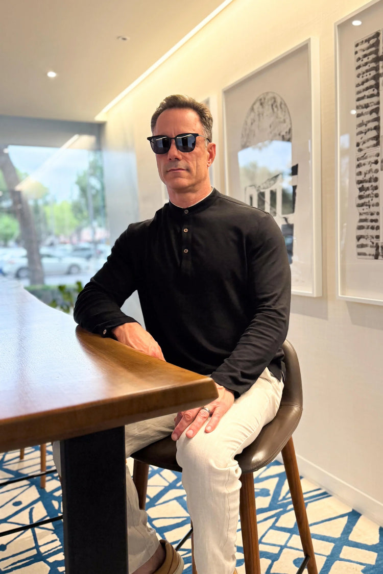 Men’s onyx black linen–Supima button-neck henley styled with linen pants, seated at a café bar for a relaxed, refined look.
